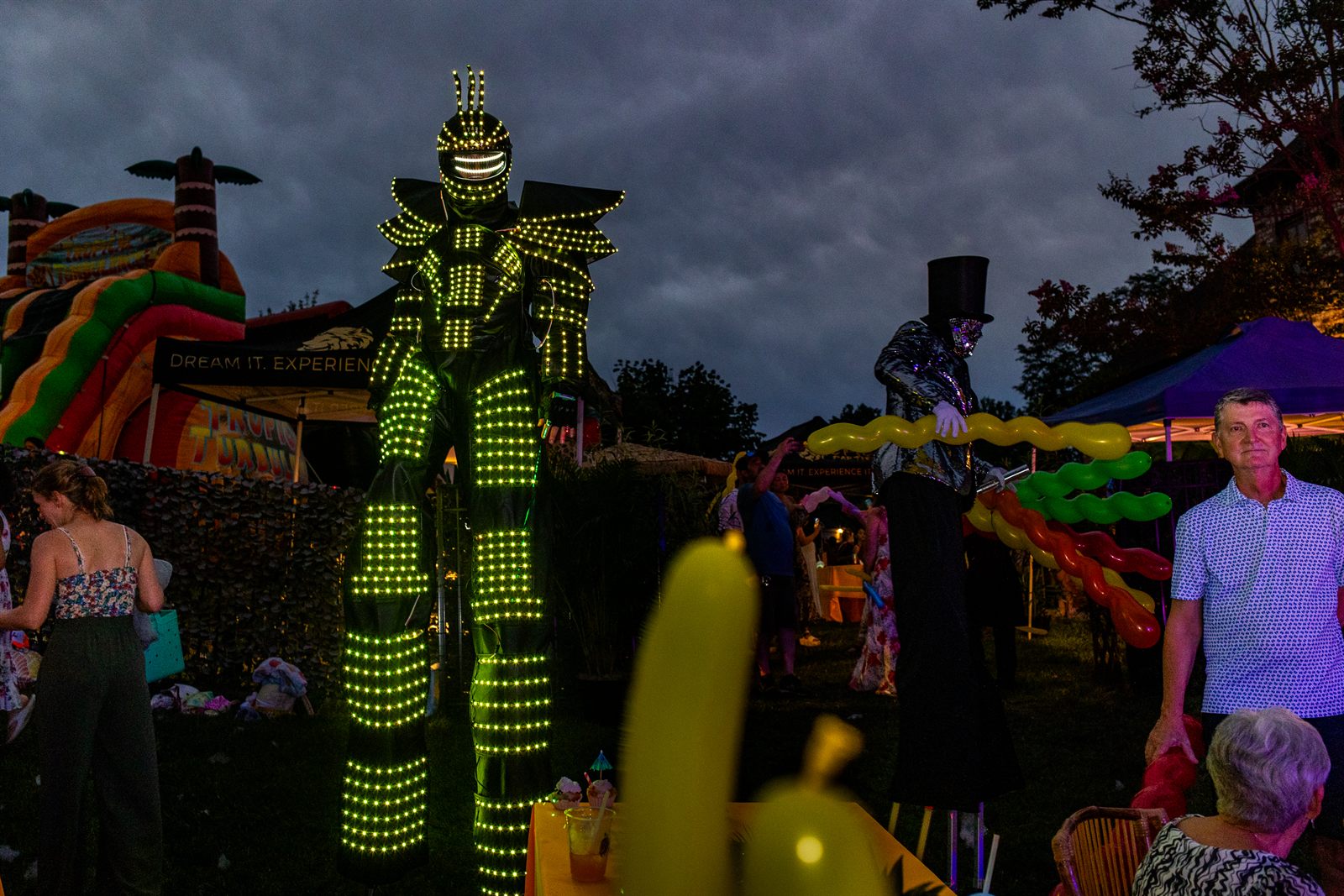 Towering LED-suit robot stilt walker glows green beside a top-hat balloon artist at dusk